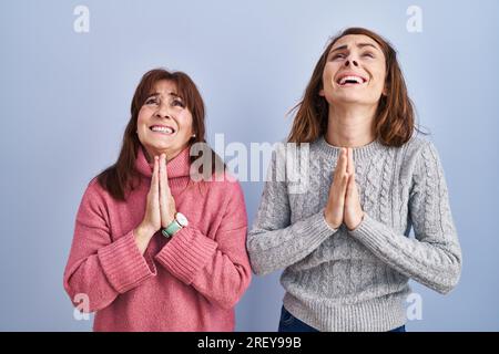 Mutter und Tochter stehen über blauem Hintergrund und betteln und beten mit Händen zusammen mit dem Ausdruck der Hoffnung im Gesicht, sehr emotional und besorgt. Betteln Stockfoto