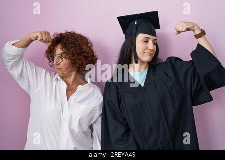 Hispanische Mutter und Tochter tragen Abschlussmütze und Morgenmantel, die zeigen, dass die Armmuskeln stolz lächeln. Fitnesskonzept. Stockfoto