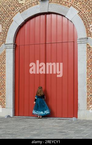 Ein kleines Mädchen in einem mittelalterlichen Kleid klopft an die großen roten Tore eines alten Schlosses. Vertikales Format Stockfoto