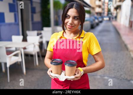 Junge schöne arabische Kellnerin lächelt selbstbewusst und hält Kaffee zum Mitnehmen auf der Terrasse des Cafés Stockfoto