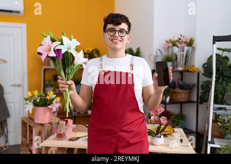 Junger nicht binärer Mann, der im Blumenladen arbeitet und dem Smartphone ein Lächeln auf dem Bildschirm zeigt, mit einem fröhlichen und kühlen Lächeln im Gesicht. Zähne zeigen. Stockfoto