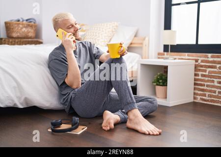 Junger weißer Mann, der mit dem Smartphone spricht und Kaffee im Schlafzimmer trinkt Stockfoto