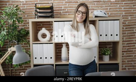Junge schwangere Geschäftsfrau, die selbstbewusst im Stehen lächelt, die Arme gekreuzt, Geste im Büro Stockfoto