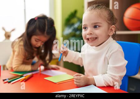 Süße Vorschulkinder, die im Kindergarten auf einem Tisch sitzen und auf Papier zeichnen Stockfoto