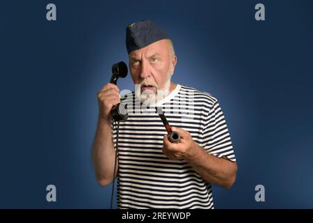 Ein echter Seemann aus der Vergangenheit spricht am Telefon. Hochformat auf blauem Hintergrund. Ein alter Seemann mit einer Rauchpfeife und einem alten Telefon in Hi Stockfoto