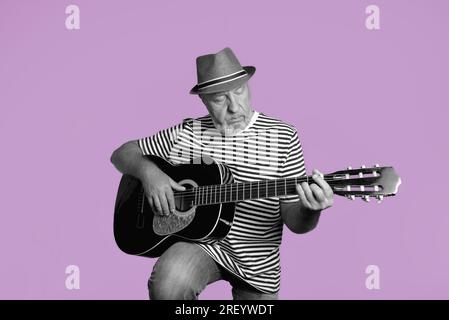 Ein älterer Mann spielt Akustikgitarre. Gestreiftes T-Shirt, Strohhut auf dunkelrosa Hintergrund. Retro-Konzept. Schwarzweiß-Porträt auf dunkelrosa Stockfoto