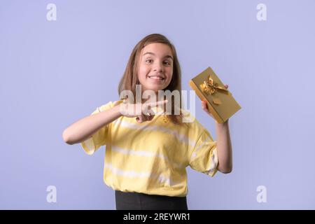 Ein fröhliches Teenager-Mädchen hat ein Geschenk in einem goldenen Paket mit einer Schleife in der Hand. Lila Hintergrund Stockfoto