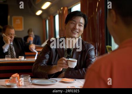 Ein Mann lächelt und hält eine Tasse Kaffee in einem Speisewagen in der Hand, der mit einem anderen Passagier interagiert. Stockfoto