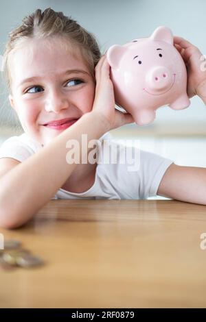 Fröhliches weißes Mädchen, das ein lächelndes rosa Sparschwein in den Händen auf einem Holztisch schüttelt. Das Sparkonzept für Kinder. Vertikaler Schuss. Stockfoto