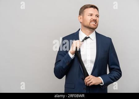 Ein müder Geschäftsmann legt Krawatten im offiziellen Anzug ab, mit einem unzufriedenen Gesicht auf grauem Hintergrund. Stockfoto