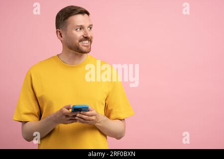 Ein fröhlicher, fröhlicher Kerl, der sein Smartphone hält und sich davon wegschaut, wenn er die leere Studiowand beiseite stellt Stockfoto