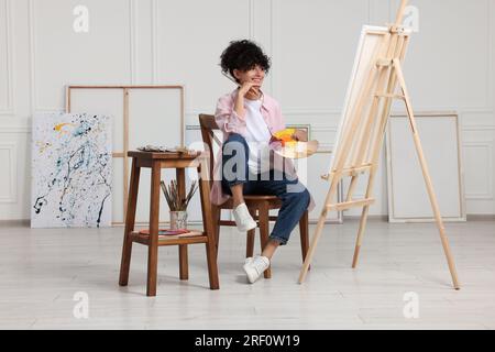 Junge Frau mit Pinsel und Künstlerpalette in der Nähe der Staffelei mit Leinwand Stockfoto