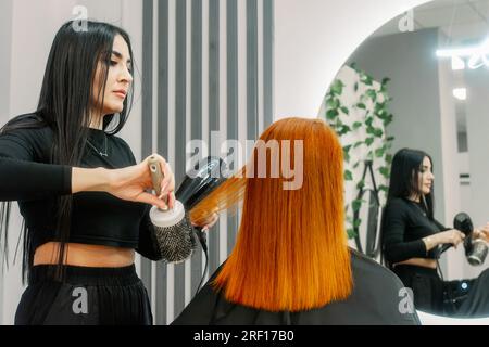 Eine Friseurin mit orientalischem Aussehen arbeitet im Schönheitssalon und macht Frisuren für Kunden. Roter Haarfärbestoff. Trockenes Haar mit Haartrockner. Schaut in den Spiegel. Sitzt auf dem Stuhl. Stockfoto