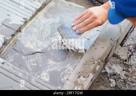 Der Baumeister verfugt Fliesen auf dem Fundament des Gebäudes mit Zement. Reparatur des toten Bereichs. Maurer legt Fliesen. Hand des Arbeiters mit Kelle-Nahaufnahme. Stockfoto