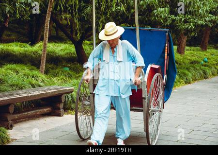 Asiatischer Seniorenmann mit blauem Anzug und Hut, der mit seinem Fahrrad im Park spaziert. Stockfoto