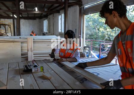 Antakya, Türkei. 18. Juli 2023. Im Rahmen des Projekts „Yuva“ (Nest) bauen Freiwillige im Bezirk Samandag ökologische Holzhäuser, die Unternehmen oder Privatpersonen über eine Partnerorganisation an Erdbebenopfer spenden können. Das Projekt „Yuva“ (Nest) beschäftigt 15 Frauen und 15 Männer aus der Region, die selbst vom Erdbeben betroffen sind. Notunterkünfte in der Hitze und den Schutt - sechs Monate nach den Erdbeben im Südosten der Türkei am 6. Februar leiden die Menschen noch immer unter den Folgen. Kredit: Bradley Secker/dpa/Alamy Live News Stockfoto