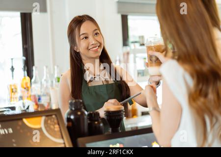 Ein asiatischer Barista überreicht Eiskaffee an weibliche Gäste über die Theke im Café. Die Kellnerin gab den Auftrag an die Gäste im Café Stockfoto
