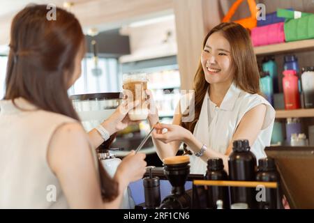 Ein asiatischer Barista überreicht Eiskaffee einer Kundin über die Theke im Café des Cafés. Eine Kundin, die das Getränk von einer Kellnerin erhält Stockfoto