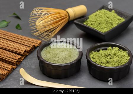 Matcha-Teepulver und in Matcha-Tee eine schwarze Schüssel. Bambusschläger und Messlöffel auf dem Tisch. Grauer Hintergrund. Stockfoto