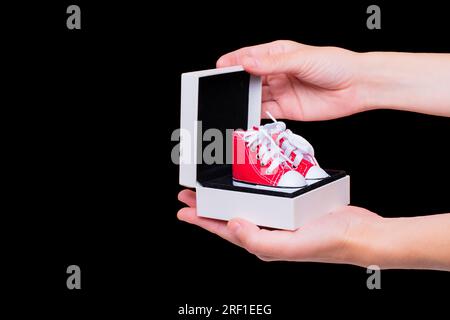 Nahaufnahme von weiblichen Händen, die eine Geschenkbox mit einem neuen Paar roter Stoffschuhe in Spielzeuggröße öffnen, isoliert auf schwarzem Hintergrund. Einkaufen, Geschenke, Feiertage Stockfoto