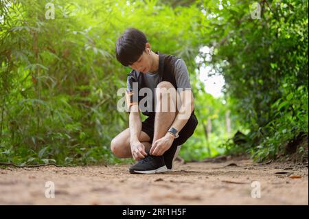 Ein sportlicher und aktiver junger asiatischer Mann in Sportbekleidung, der sich beim Laufen im grünen Wald die Schnürsenkel bindet. Sommeraktivität, Sport, Lifestyle Stockfoto