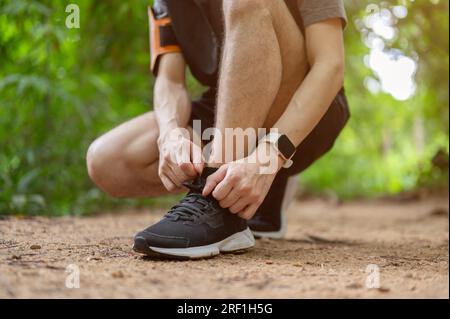 Nahaufnahme eines sportlichen und aktiven jungen asiatischen Mannes in Sportbekleidung, der sich beim Laufen im grünen Wald die Schnürsenkel bindet. Stockfoto