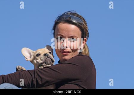Junge blonde Frau, die eine 12 Wochen alte französische Bulldogge in den Armen hält. Blauer Himmel im Hintergrund Weitere Motive zu diesem Thema sind auf Anfrage erhältlich Stockfoto