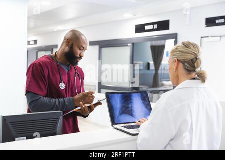 Verschiedene männliche und weibliche Ärzte diskutieren über Arbeit und verwenden Laptop in der Rezeption des Krankenhauses Stockfoto