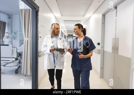 Glückliche weisse Ärztinnen mit Stethoskopen, die Tabletten im Flur des Krankenhauses benutzen Stockfoto