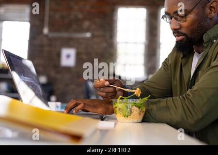 Ein afroamerikanischer Geschäftsmann, der ein Notebook benutzt und Salat zum Mitnehmen am Schreibtisch im Büro isst Stockfoto