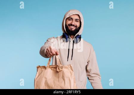 Fröhlicher, lächelnder araber in Kapuze, der eine Papiertüte mit Porträt zum Mitnehmen gibt. Restaurantzustelldienst, Kurierdienst, der das Paket mit Mittagessen zum Mitnehmen bereithält und in die Kamera schaut Stockfoto