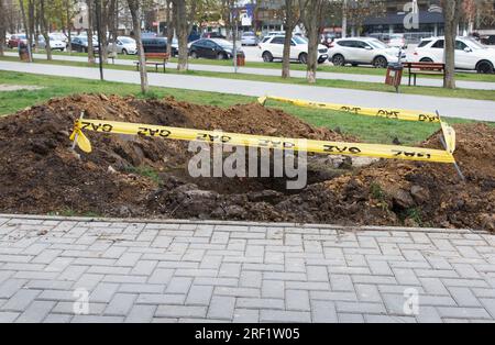 Eine quadratische Grube, die in den Boden gegraben wurde, neben dem öffentlichen Bürgersteig und eingezäunt mit einem gelben Band, als Zeichen der Gefahr. Kommunale Dienstleistungen, Reparatur von UN Stockfoto