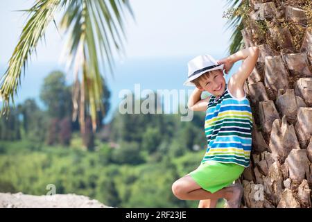 Ein glücklicher Junge in einem panama steht neben einer tropischen Palme, sieht in die Kamera und lächelt. Ein Kind im Urlaub in einem tropischen Land. Stockfoto