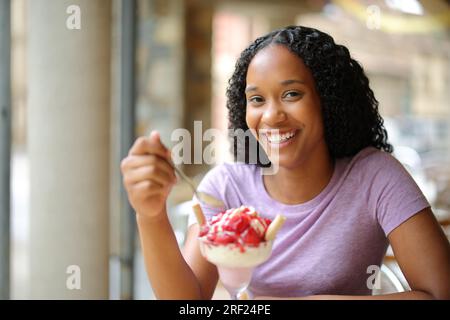 Glückliche schwarze Frau, die Dessert auf einer Restaurantterrasse isst und vor die Kamera schaut Stockfoto