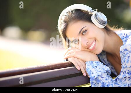 Wunderschönes Porträt einer Frau, die Kopfhörer trägt, auf einer Bank im Park, die dich ansieht Stockfoto