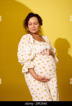 Authentisches Porträt einer schönen schwangeren Frau in einem weißen, stilvollen Sommerkleid, die ihren großen Bauch berührt, lächelnd zur Seite schaut, isolierter gelber Hengst Stockfoto