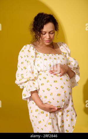 Multiethnische lockige Frau mittleren Alters schöne schwangere Frau, die Hände auf den Bauch legt und Babykicks genießt, isoliert auf gelbem Hintergrund. Pregna Stockfoto