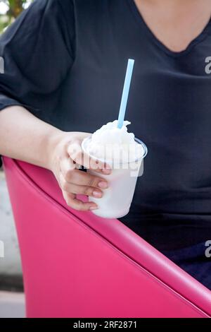 Eine Frau, die eine Kokosnuss-Slush in einem Plastikbecher hält, sitzt auf einem pinkfarbenen Sofa. Stockfoto