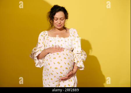 Eine charmante schwangere Frau, die ihren großen Bauch hält, im Sommerkleid angezogen ist, mit geschlossenen Augen über gelbem Studiohintergrund posiert und kostenlose Werbung macht Stockfoto