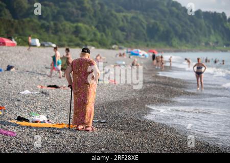 04. August 2022. Georgien, Batumi. Strand. Eine ältere Frau mit einem Stock ruht auf einem felsigen Ufer. Stockfoto