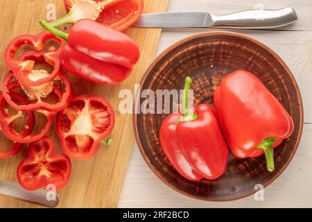 Mehrere rote Paprika im Ganzen und in Scheiben geschnitten mit Tonplatte und Messer auf Holztisch, Makro, Draufsicht. Stockfoto