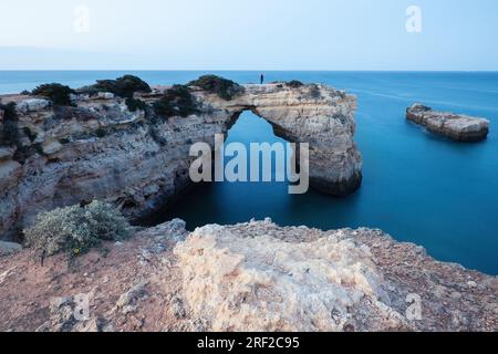 Eine Frau steht am natürlichen Bogen am Marinha-Strand, Algarve Portugal. Felsenklippen am Marinha-Strand und türkisfarbenes Meerwasser an der Küste Portugals in der Algarve. Stockfoto