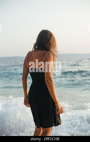 Rückansicht einer jungen Frau in schwarzem Kleid, die im Meer seicht steht Stockfoto