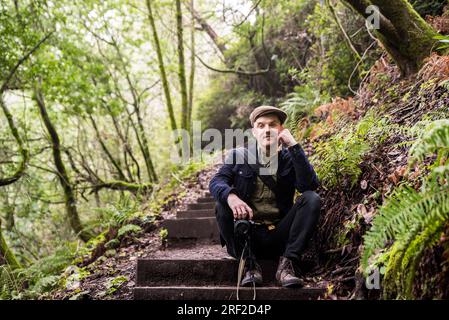 Ein Mann, der vor der Kamera lächelt und auf einer hölzernen Treppe im Wald sitzt Stockfoto