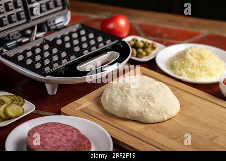 Pizza-Waffel kochen, Piffle. Waffeleisen und Hefeteig für Pizza und verschiedene Zutaten - Salami, Käse, Oliven, Tomaten auf dem Küchentisch. Stockfoto