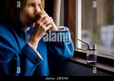 Eine Frau trinkt Kaffee und raucht Cannabis. Eine Erwachsene Frau über 40 sitzt neben dem Fenster, daneben befindet sich ein Gerät zum Rauchen von Marihuana. Stockfoto