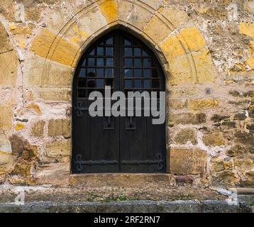 Alte dunkle Holztür mit spitzem Bogen in der Steinmauer, in der kantabrischen Stadt Santillana del Mar. Stockfoto