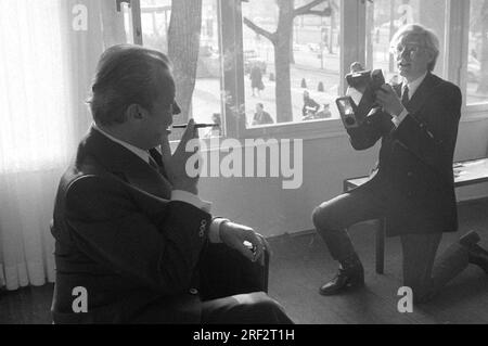 ARCHIVFOTO: Andy WARHOL wäre am 6. August 2023 95 Jahre alt gewesen, ART/Politics Andy WARHOL(r.), USA, Künstler, Darstellung von Ex-Kanzler Willy BRANDT (SPD) mit einer Sofortkamera. Warhol kniet auf einem Bein und hält die Kamera aufrecht, schaut aber nicht durch sie, sondern scheint Brandt Anweisungen zu geben. Brandt sitzt auf einem Stuhl und hält eine Zigarette mit einem Mundstück in der rechten Hand, halblang im Profil und ziemlich Silhouettenartig, aber immer noch erkennbar. Warhol ist von vorne in voller Länge zu sehen. Schwarzweißaufnahme im Querformat. ? Stockfoto