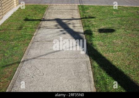 Betonpfad im Westen Sydneys mit Grasstreifen auf beiden Seiten und dem Schatten eines hölzernen Stabs und Drähten, die den Weg überqueren Stockfoto