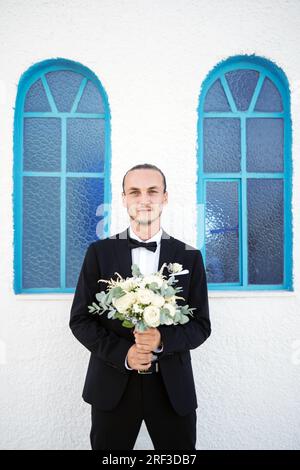 Stilvoller Bräutigam wartet vor der Kirche auf die Braut mit einem Blumenstrauß Stockfoto